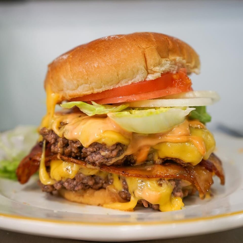 A towering double cheeseburger with bacon, lettuce, tomato, onion, and special sauce on a white plate.