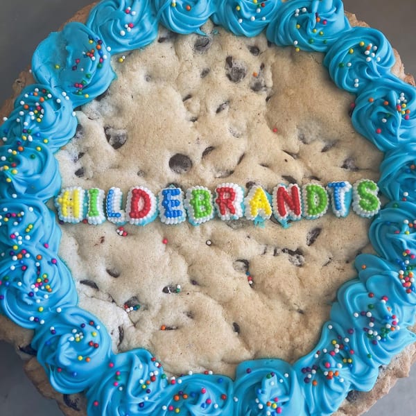 A giant chocolate chip cookie cake with blue frosting and sprinkles, spelling "HILDEBRANDTS" in colorful letters.