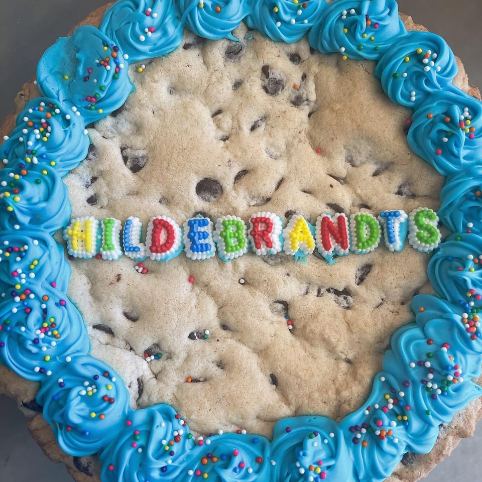 A giant chocolate chip cookie cake with blue frosting and sprinkles, spelling "HILDEBRANDTS" in colorful letters.